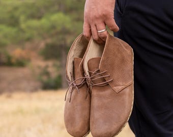 May include: A pair of brown leather ankle boots with laces. The boots have a rounded toe and a stitched sole. The leather has a textured appearance. The boots are held in a person's hand.