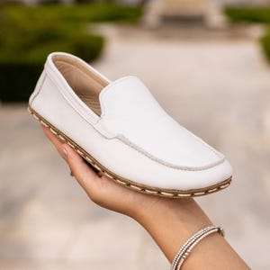 Op de afbeelding: Witte leren loafer met een ronde neus en een lichtbruine zool. De schoen wordt in een hand gehouden, wat het strakke design en de minimalistische esthetiek laat zien. De zool lijkt gestikt met een contrasterende draad.