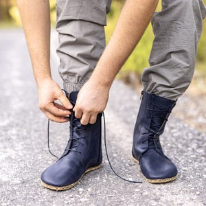 May include: Navy blue leather boots with a lace-up design. The boots have a natural-colored sole and are being laced up. The person is wearing gray pants. The boots appear to be designed for outdoor activities.