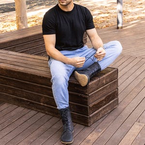 May include: A person wearing a black t-shirt and light blue jeans, sitting on a wooden bench. The person is wearing black lace-up boots with a tan sole. The boots appear to be made of suede or a similar material.