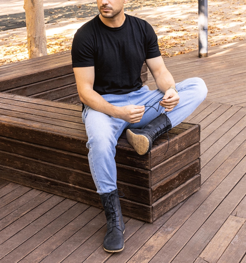 May include: A person wearing a black t-shirt and light blue jeans, sitting on a wooden bench. The person is wearing black lace-up boots with a tan sole. The boots appear to be made of suede or a similar material.
