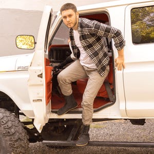 May include: A person exiting a white pickup truck with a red interior. The person is wearing a white t-shirt, gray pants, and black boots. They are also wearing a black and white plaid shirt.