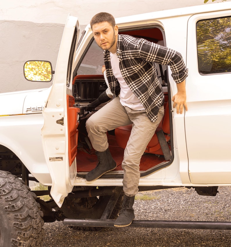 May include: A person exiting a white pickup truck with a red interior. The person is wearing a white t-shirt, gray pants, and black boots. They are also wearing a black and white plaid shirt.