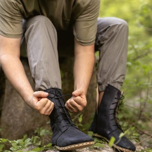 May include: Black leather boots with laces. The boots have a high-top design and are being laced up. The boots are worn outdoors, with a natural background of green foliage.