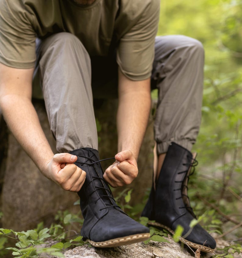 May include: Black leather boots with laces. The boots have a high-top design and are being laced up. The boots are worn outdoors, with a natural background of green foliage.