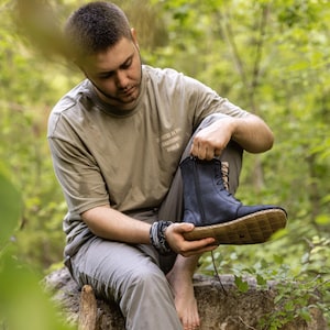 May include: A person holding a black leather boot with a side zipper and lace-up closure. The boot has a light brown sole with a diamond pattern. The person is wearing a tan t-shirt and gray pants.