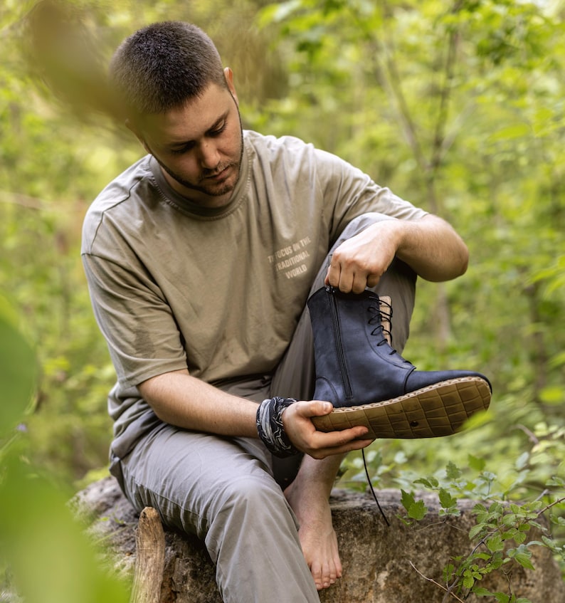 May include: A person holding a black leather boot with a side zipper and lace-up closure. The boot has a light brown sole with a diamond pattern. The person is wearing a tan t-shirt and gray pants.