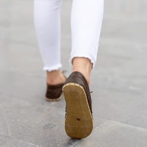May include: Close-up of brown leather moccasin-style shoes with a light-colored sole. The shoes are being worn with white jeans with a frayed hem. The image is taken from a low angle, showing the sole of the shoe.