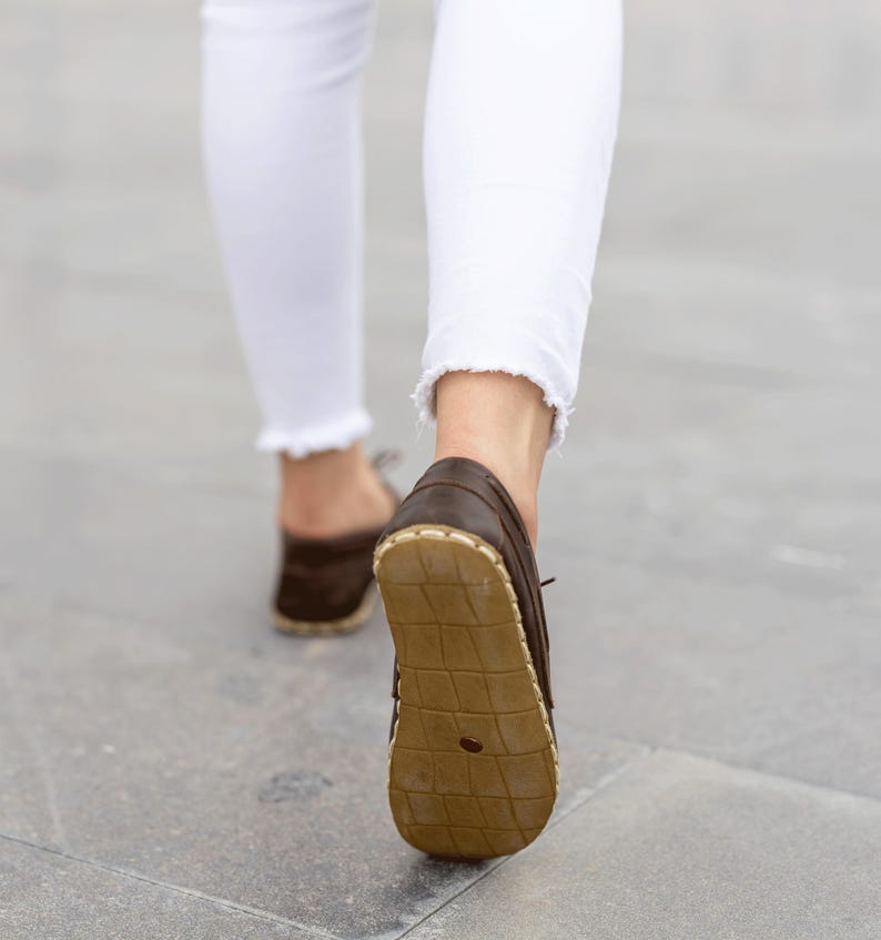 May include: Close-up of brown leather moccasin-style shoes with a light-colored sole. The shoes are being worn with white jeans with a frayed hem. The image is taken from a low angle, showing the sole of the shoe.
