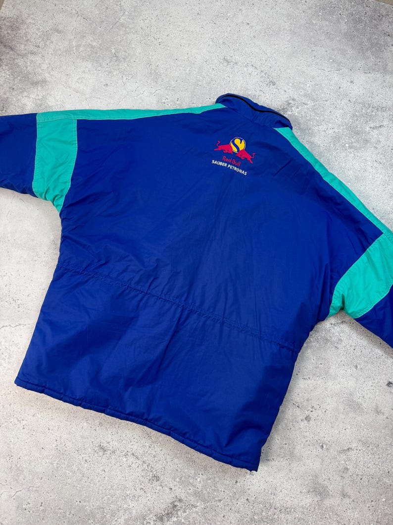 Pu&ograve; includere: Giacca blu e verde acqua con il logo Red Bull Sauber Petronas sul retro. La giacca ha un corpo blu con accenti verde acqua sulle maniche e sulle spalle. Il logo Red Bull &egrave; rosso e giallo.