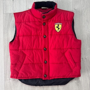 Könnte beinhalten: Eine rote Steppweste mit schwarzem Kragen und Futter. Die Weste hat eine Knopfleiste, zwei Taschen und einen Ferrari-Logoaufnäher auf der Brust. Die ärmellose Weste scheint aus leichtem Material gefertigt zu sein.