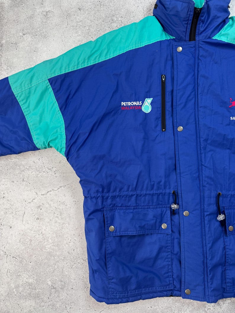 Pu&ograve; includere: Giacca vintage blu e turchese con il logo "PETRONAS MALAYSIA". La giacca ha un cappuccio, una cerniera e pi&ugrave; tasche. Le maniche sono turchesi e il corpo blu. Ha una vita regolabile e chiusure a scatto.