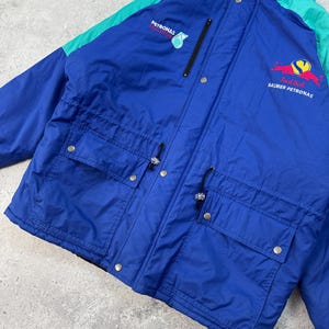 Pu&ograve; includere: Giacca vintage blu e verde acqua con il logo Red Bull Sauber Petronas sul petto. La giacca ha due tasche frontali, una chiusura con zip e il logo Petronas sulla parte superiore sinistra del petto. Realizzata in materiale leggero.