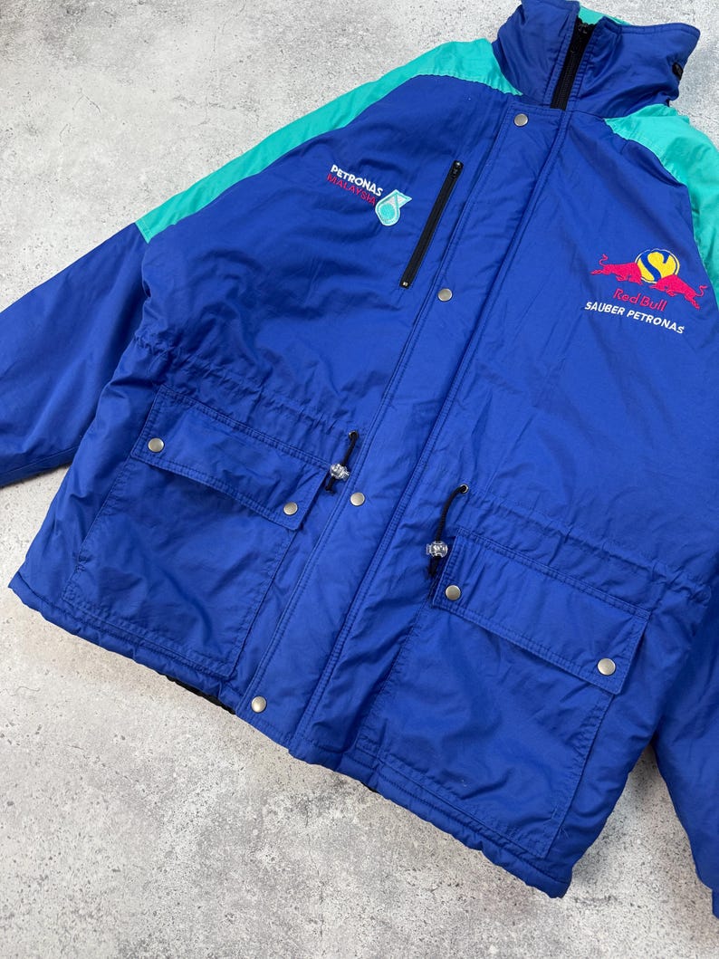 Pu&ograve; includere: Giacca vintage blu e verde acqua con il logo Red Bull Sauber Petronas sul petto. La giacca ha due tasche frontali, una chiusura con zip e il logo Petronas sulla parte superiore sinistra del petto. Realizzata in materiale leggero.