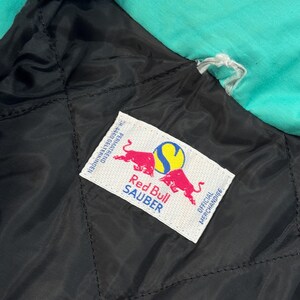 Pu&ograve; includere: Primo piano della fodera trapuntata nera di una giacca con uno strato esterno turchese. Un'etichetta bianca presenta il logo Red Bull Sauber con due tori rossi e il testo "Red Bull Sauber" e "Official Merchandise".
