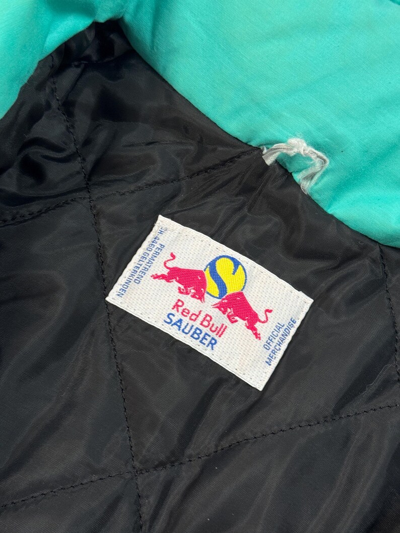Pu&ograve; includere: Primo piano della fodera trapuntata nera di una giacca con uno strato esterno turchese. Un'etichetta bianca presenta il logo Red Bull Sauber con due tori rossi e il testo "Red Bull Sauber" e "Official Merchandise".