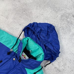 Pu&ograve; includere: Giacca con cappuccio blu, verde acqua e viola. La giacca ha un cappuccio con coulisse e un interno verde acqua. La giacca ha una chiusura con bottoni e un logo sul colletto interno. La giacca &egrave; realizzata in materiale leggero.