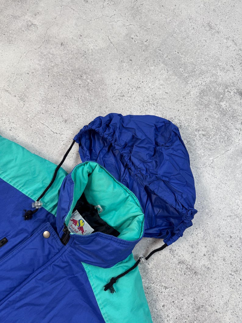 Pu&ograve; includere: Giacca con cappuccio blu, verde acqua e viola. La giacca ha un cappuccio con coulisse e un interno verde acqua. La giacca ha una chiusura con bottoni e un logo sul colletto interno. La giacca &egrave; realizzata in materiale leggero.