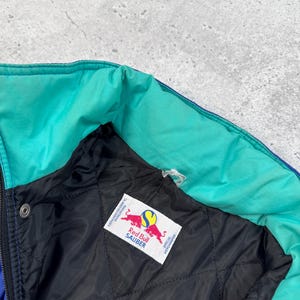 Pu&ograve; includere: Primo piano di una giacca multicolore con colletto turchese e interno trapuntato nero. La giacca presenta un'etichetta Red Bull Sauber. La giacca &egrave; blu, turchese e nera.