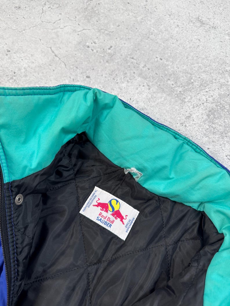 Pu&ograve; includere: Primo piano di una giacca multicolore con colletto turchese e interno trapuntato nero. La giacca presenta un'etichetta Red Bull Sauber. La giacca &egrave; blu, turchese e nera.