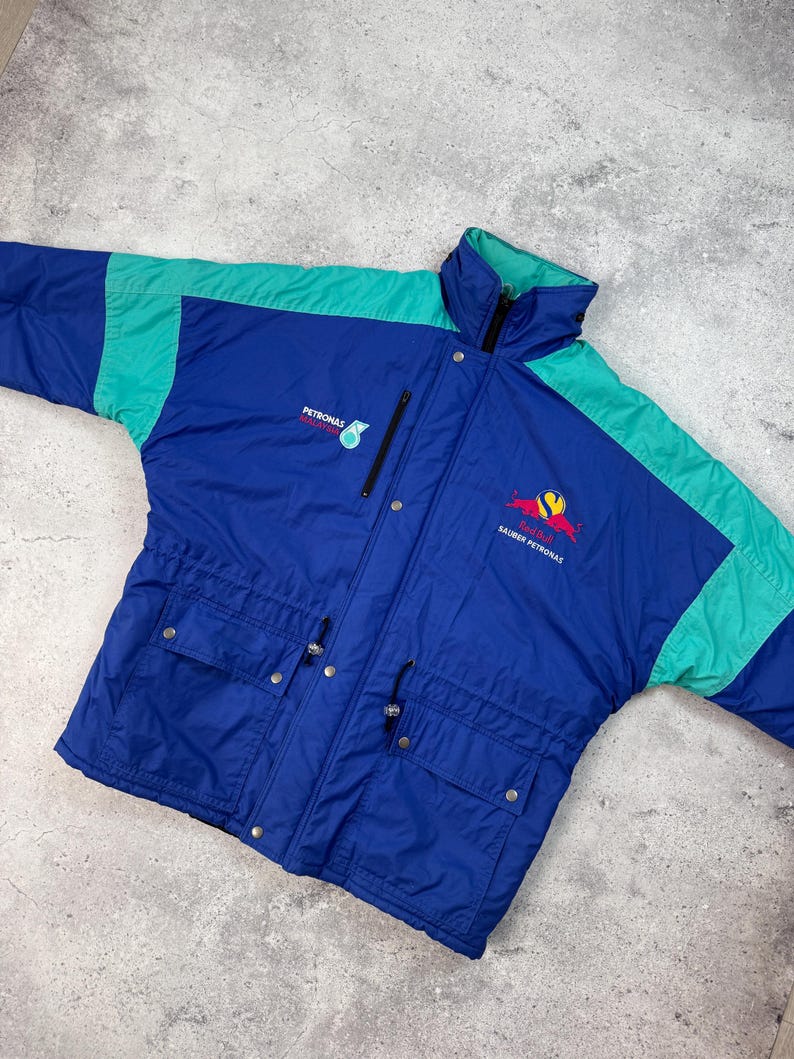 Pu&ograve; includere: Giacca vintage blu e verde acqua con i loghi "PETRONAS" e "SAUBER PETRONAS". La giacca ha un collo alto, una cerniera e due tasche frontali. La giacca &egrave; realizzata in materiale leggero.