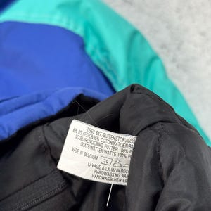 Pu&ograve; includere: Primo piano di una giacca multicolore con sezioni blu, verde acqua e nere. Un'etichetta con testo in pi&ugrave; lingue &egrave; visibile, tra cui "MADE IN BELGIUM" e istruzioni per il lavaggio.