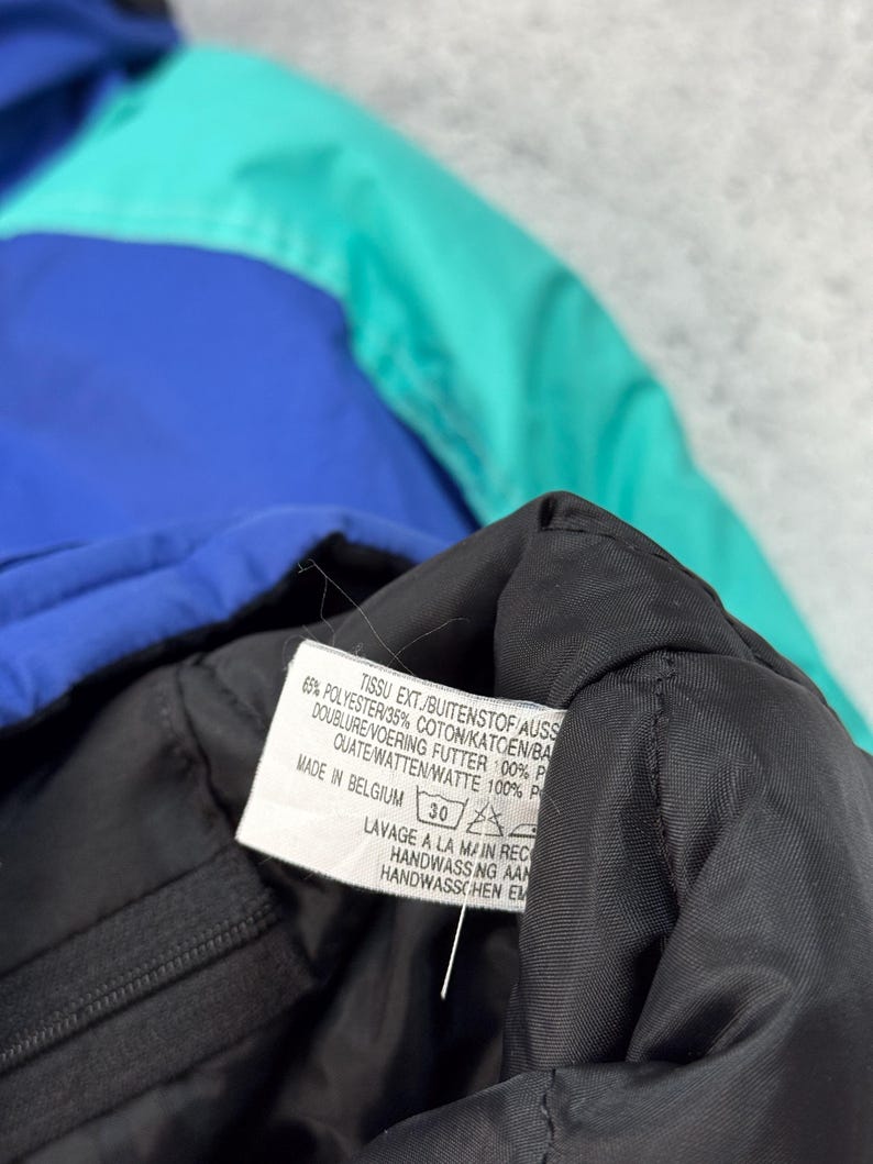 Pu&ograve; includere: Primo piano di una giacca multicolore con sezioni blu, verde acqua e nere. Un'etichetta con testo in pi&ugrave; lingue &egrave; visibile, tra cui "MADE IN BELGIUM" e istruzioni per il lavaggio.