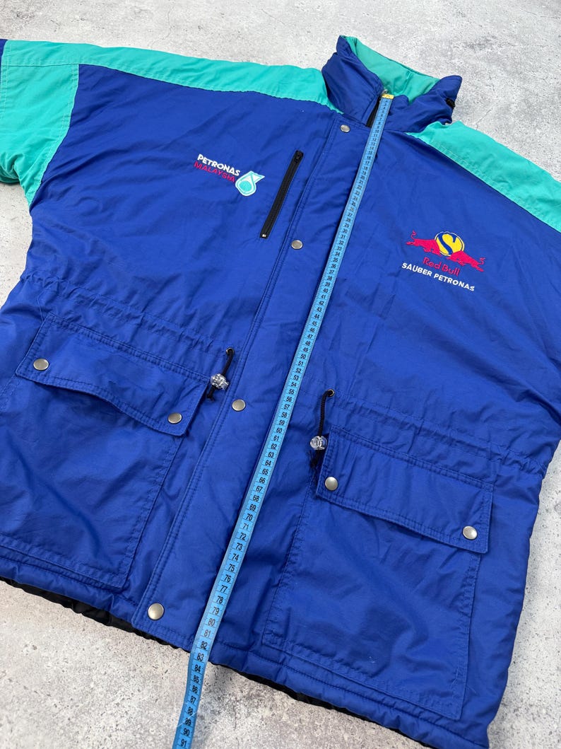 Pu&ograve; includere: Giacca vintage blu e verde acqua con i loghi Petronas e Red Bull Sauber Petronas. La giacca ha una chiusura a zip, due tasche frontali e una vita con coulisse. Un metro a nastro &egrave; appoggiato sulla giacca.