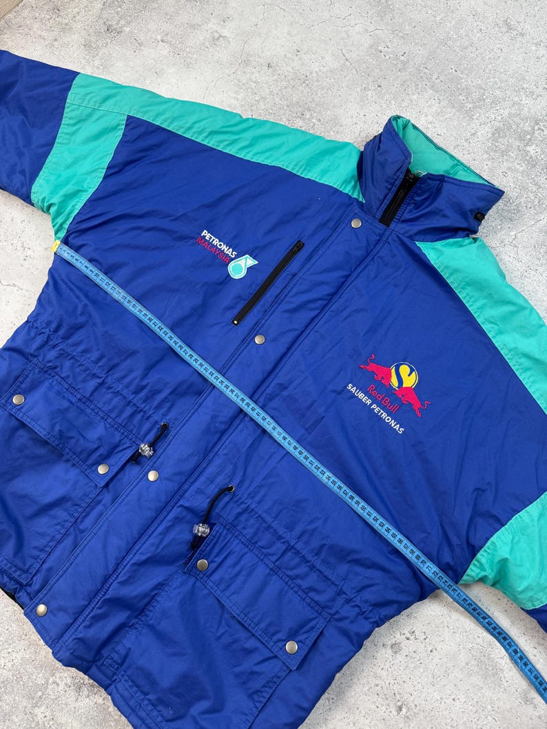 Pu&ograve; includere: Giacca vintage blu e verde acqua con i loghi Petronas e Red Bull Sauber Petronas. La giacca ha una chiusura a zip, tasche multiple e un cordoncino in vita. Un metro a nastro &egrave; appoggiato sulla giacca.