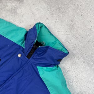 Pu&ograve; includere: Giacca blu, verde acqua e nera con chiusura a zip e bottone a pressione. La giacca ha un collo alto e una fodera verde acqua. La giacca &egrave; realizzata in un materiale lucido.