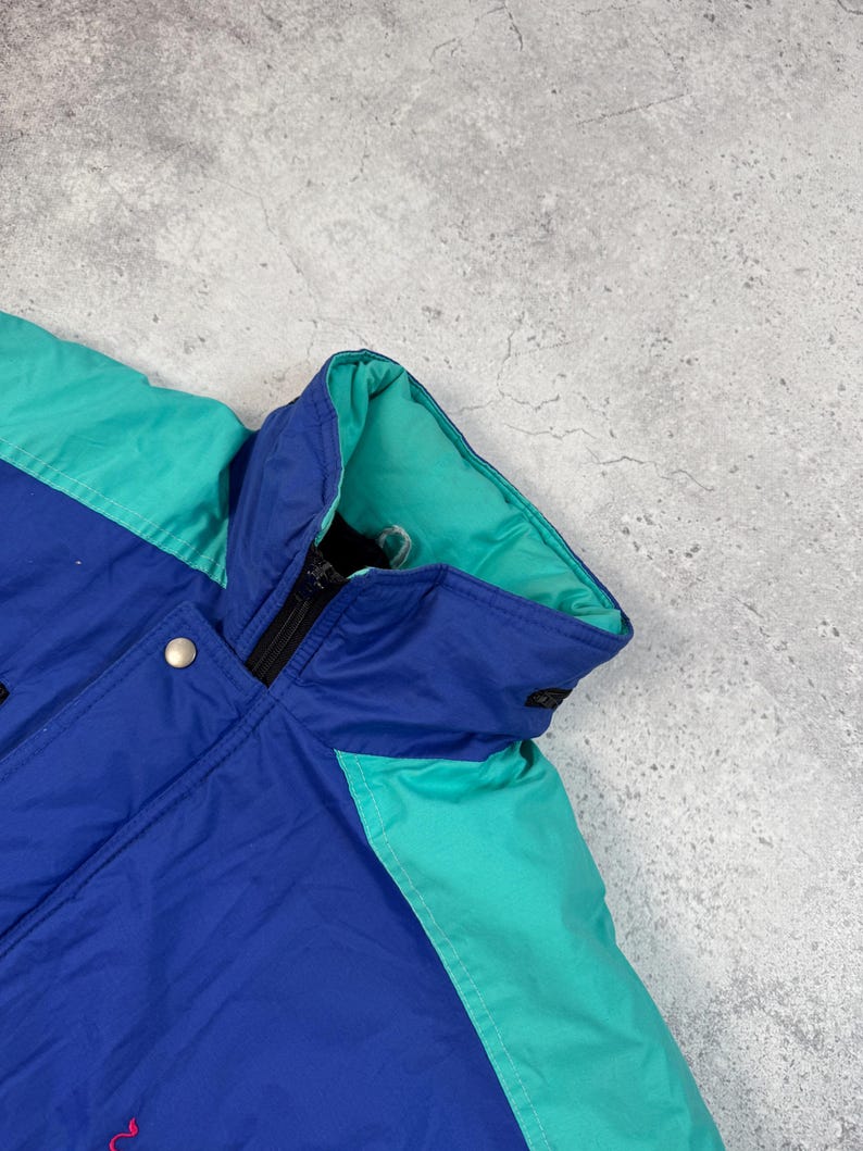Pu&ograve; includere: Giacca blu, verde acqua e nera con chiusura a zip e bottone a pressione. La giacca ha un collo alto e una fodera verde acqua. La giacca &egrave; realizzata in un materiale lucido.