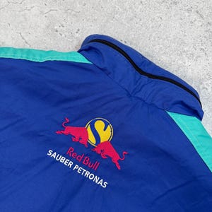Pu&ograve; includere: Giacca blu e verde acqua con il logo Red Bull Sauber Petronas ricamato sul retro. Il logo presenta due tori rossi contrapposti con un cerchio giallo e la lettera "S".