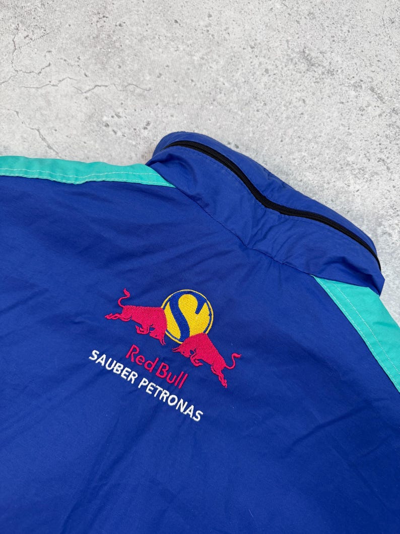 Pu&ograve; includere: Giacca blu e verde acqua con il logo Red Bull Sauber Petronas ricamato sul retro. Il logo presenta due tori rossi contrapposti con un cerchio giallo e la lettera "S".