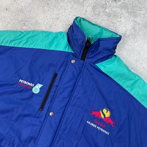 Pu&ograve; includere: Giacca da corsa blu, verde acqua e nera con il logo Red Bull Sauber Petronas. La giacca ha un collo alto, una cerniera e chiusure a scatto. &Egrave; visibile anche il logo Petronas Malaysia.