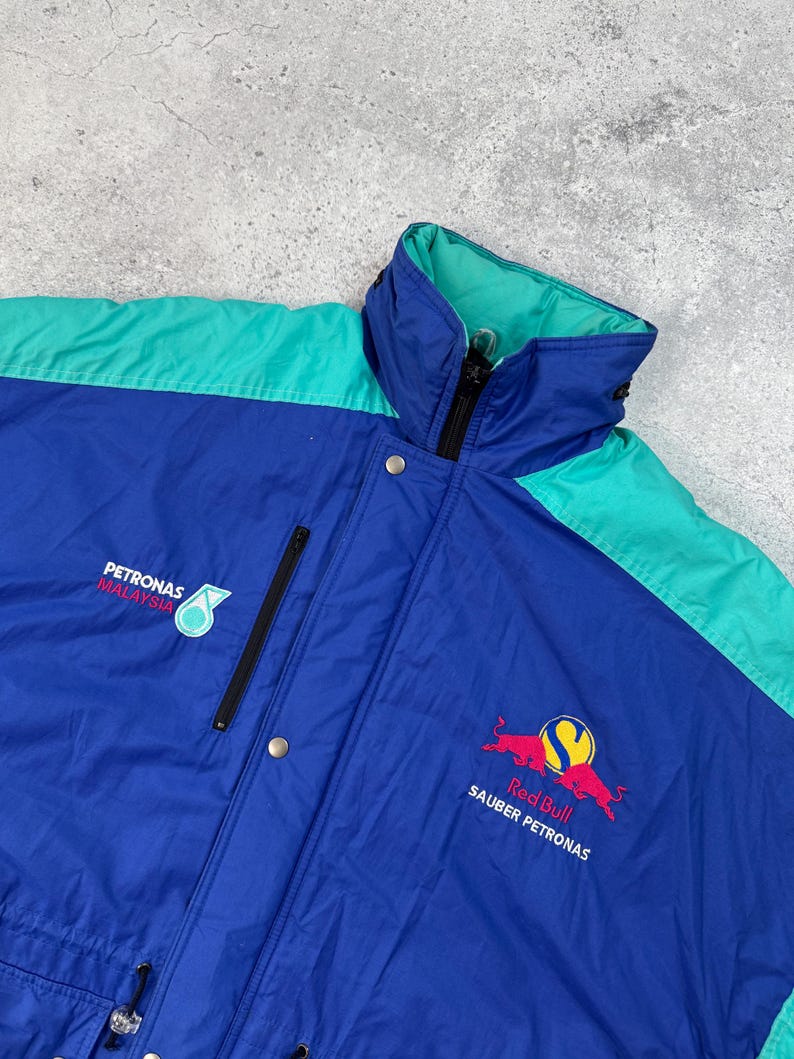 Pu&ograve; includere: Giacca da corsa blu, verde acqua e nera con il logo Red Bull Sauber Petronas. La giacca ha un collo alto, una cerniera e chiusure a scatto. &Egrave; visibile anche il logo Petronas Malaysia.