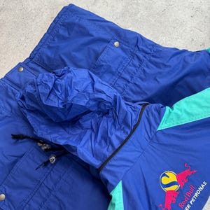 Pu&ograve; includere: Giacca vintage blu e verde acqua con cappuccio. La giacca presenta il logo Red Bull Sauber Petronas ricamato sul davanti. La giacca ha due tasche e un cappuccio con coulisse.