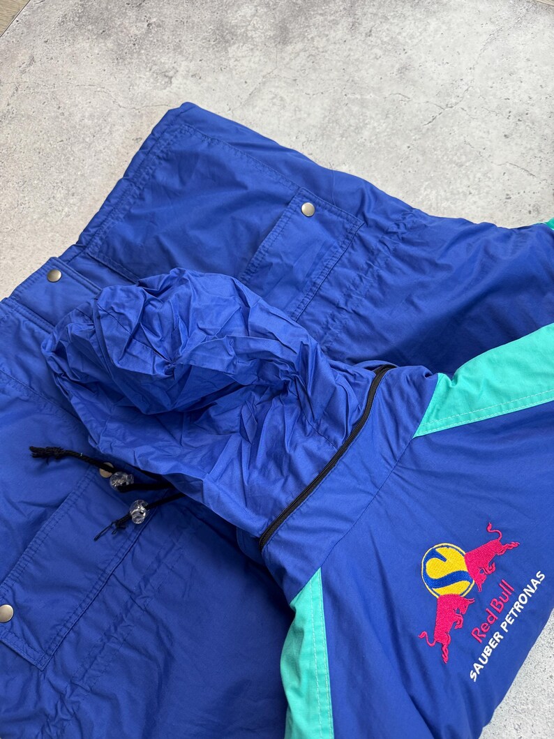 Pu&ograve; includere: Giacca vintage blu e verde acqua con cappuccio. La giacca presenta il logo Red Bull Sauber Petronas ricamato sul davanti. La giacca ha due tasche e un cappuccio con coulisse.