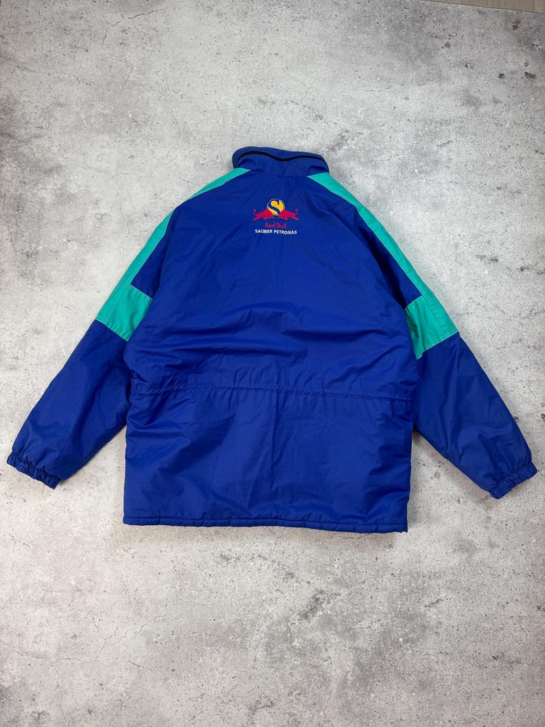 Pu&ograve; includere: Giacca blu e verde acqua con logo e testo sul retro. La giacca ha una chiusura a zip e un collo alto. Il logo &egrave; rosso e giallo. Il testo sul retro della giacca recita "SAUBER PETRONAS".