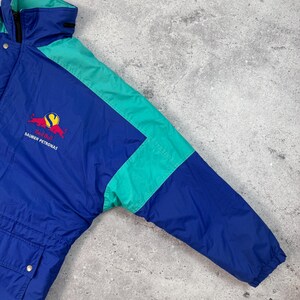 Pu&ograve; includere: Giacca Red Bull Sauber Petronas blu e verde acqua. La giacca presenta un corpo blu con accenti verde acqua sulle maniche e sul colletto. Il logo Red Bull e il testo Sauber Petronas sono visibili sul petto.