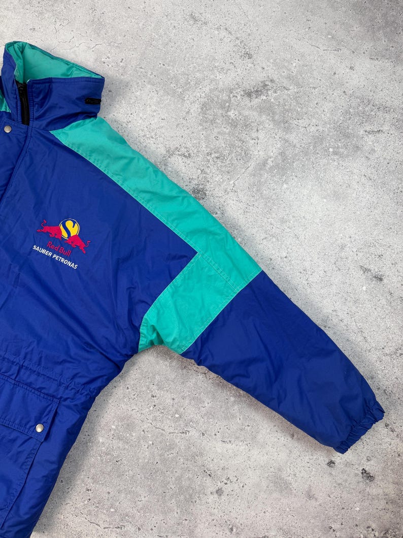 Pu&ograve; includere: Giacca Red Bull Sauber Petronas blu e verde acqua. La giacca presenta un corpo blu con accenti verde acqua sulle maniche e sul colletto. Il logo Red Bull e il testo Sauber Petronas sono visibili sul petto.