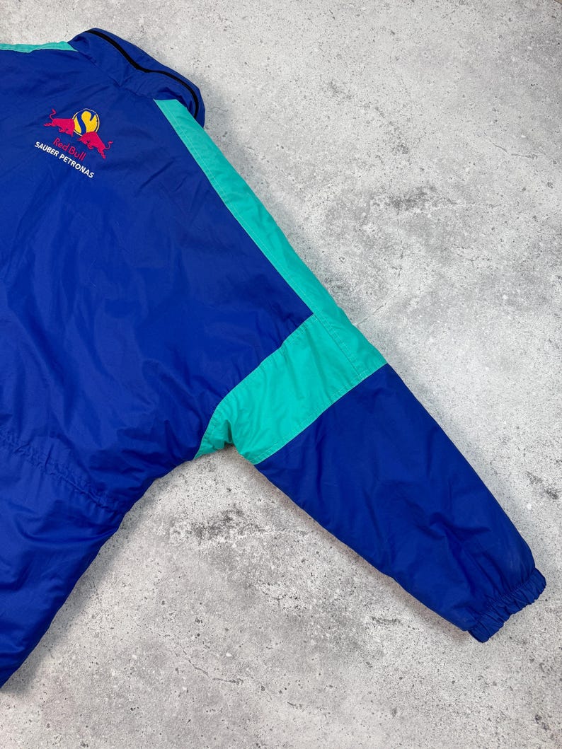 Pu&ograve; includere: Giacca vintage blu e verde acqua con logo Red Bull Sauber Petronas. La giacca presenta un design a blocchi di colore con dettagli verde acqua sulle maniche e un colletto nero. Realizzata in materiale leggero.