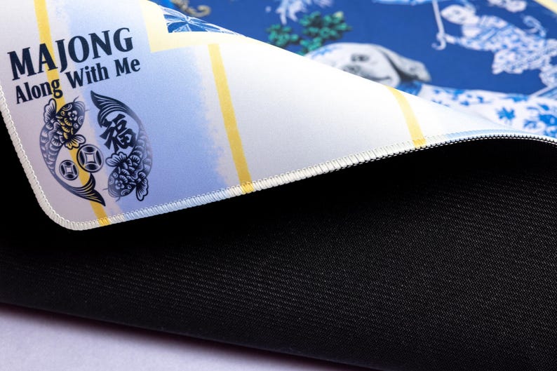 May include: A blue and white patterned mousepad with the text "MAJONG Along With Me" and a koi fish design. The mousepad has a black, textured bottom and a white stitched edge. The design includes yellow stripes.