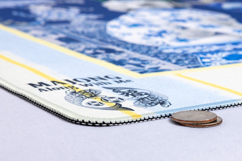 May include: A rectangular mousepad with a blue and white design, featuring text that reads "MAJONG Along With Me." The mousepad has a black stitched edge and a yellow stripe. Two coins are visible in the foreground.