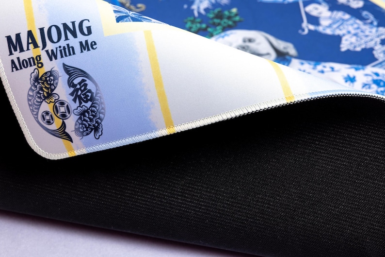 May include: A blue and white patterned mousepad with the text "MAJONG Along With Me" and a koi fish design. The mousepad has a black, textured bottom and a white stitched edge. The design includes yellow stripes.