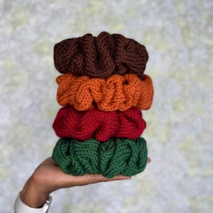 Peut inclure: Une pile de quatre chouchous tricotés aux couleurs automnales : marron, orange, rouge et vert. Les chouchous sont faits d'un fil épais et texturé et sont empilés. Le fond est un motif floral blanc et doux.