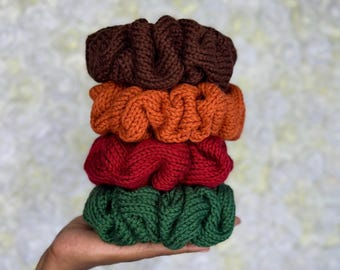 May include: A stack of four knitted scrunchies in fall colors: brown, orange, red, and green. The scrunchies are made of a thick, textured yarn and are stacked on top of each other. The background is a soft, white floral pattern.