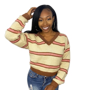 May include: Cream-colored, striped sweater with pink and brown accents. The V-neck sweater features horizontal pink stripes and brown trim on the neckline, cuffs, and hem. The sweater is made of a knitted material.