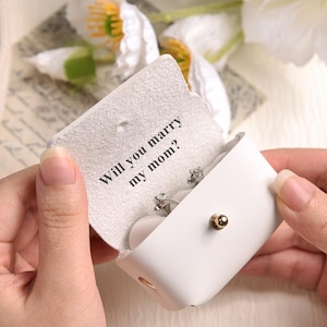 May include: A white leatherette jewelry box with a gold-colored clasp is opened to reveal a ring. The inside of the box has the text "Will you marry my mom?" printed on it. The box is held by two hands.