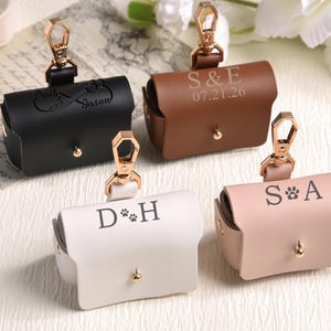 Könnte beinhalten: Vier personalisierte Hundekotbeutelhalter aus Leder in Schwarz, Braun, Weiß und Rosa. Jeder Halter hat einen goldfarbenen Verschluss und einen kleinen goldfarbenen Knopf. Die Halter sind mit Initialen, Namen und einem Datum graviert.