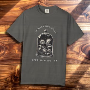 May include: Dark gray t-shirt featuring a detailed illustration of a Venus flytrap plant inside a glass cloche. The text "Dionaea Muscipula" and "Specimen No. 47" are printed above and below the illustration. The shirt is displayed on a wooden surface.